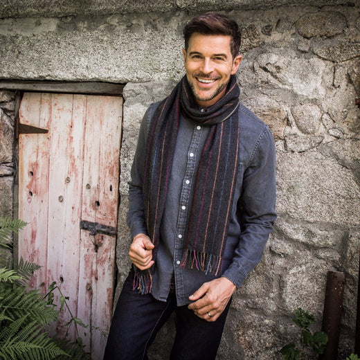 A man standing in front of a stone wall wearing a charcoal gray scarf with multicolored pinstripes.