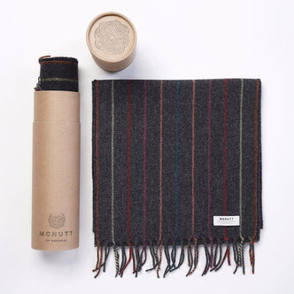 Charcoal McNutt scarf with multicolored vertical stripes and fringe, shown folded beside its cylindrical kraft packaging.