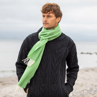 Green herringbone lambswool scarf with cream fringe, worn over a dark cable‑knit sweater on a cloudy beachside day.