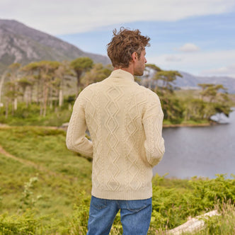 A man stands with his back to the camera, wearing a cream-colored cable-knit aran sweater and blue jeans. He overlooks a serene landscape featuring a lake surrounded by lush greenery and mountains in the background under a clear sky.