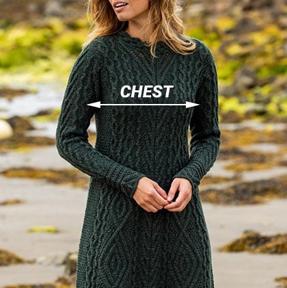 A woman stands on a beach wearing a dark green knit aran sweater dress. The dress features a textured pattern, and an arrow labeled 