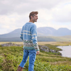 A man wearing a hand-knitted blue sweater with a white sheep pattern standing in a field.