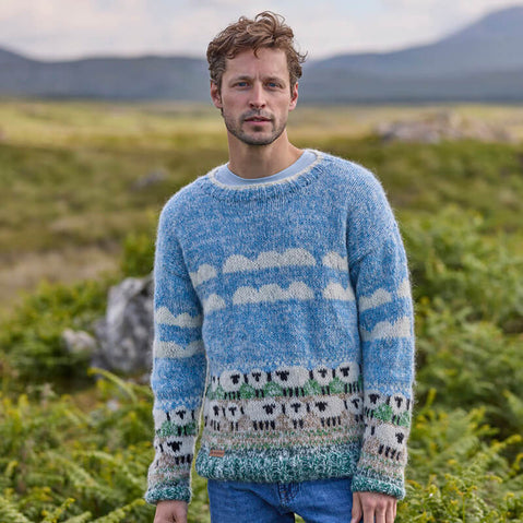 A man wearing a hand-knitted blue sweater with a white sheep pattern standing in a field.