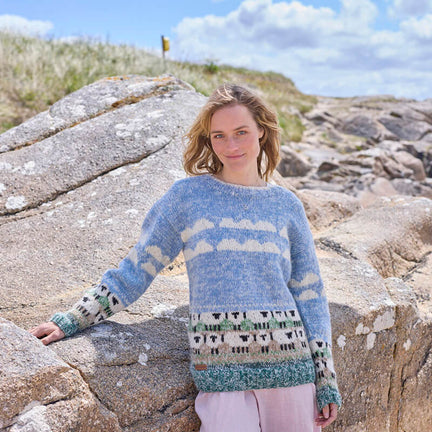 A woman standing by rock wearing a hand-knitted grey and white sheep patterned sweater.