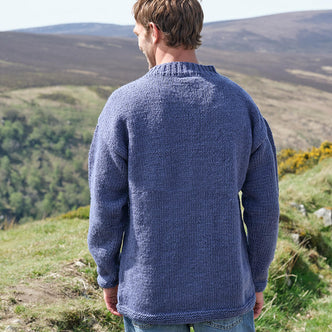 Man wearing a blue knitted sweater standing on a grassy hill with a scenic view.