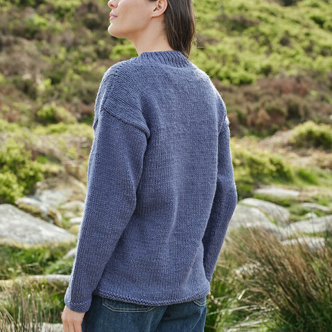 Person wearing a blue Aran sweater standing in a natural setting with greenery and rocks.
