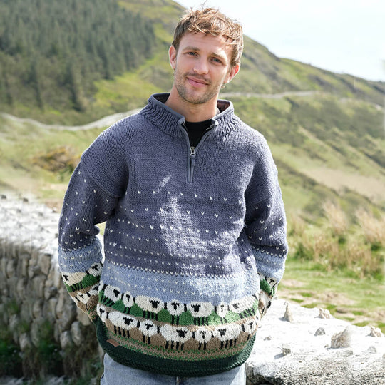 Man wearing a patterned sheep sweater standing in a natural setting with mountains and sheep.