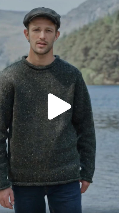 Person standing outdoors wearing a textured knit sweater and a flat cap, positioned beside a lake with wooded hills in the background.