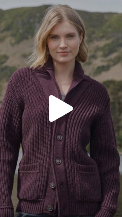 Person standing outdoors wearing a textured knitted shawl‑collar cardigan with front buttons, photographed against a green hillside landscape.