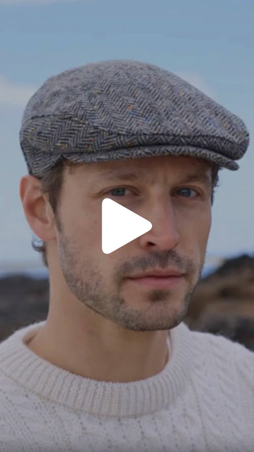 Close‑up of a person wearing a gray herringbone flat cap, with a blurred outdoor landscape in the background.