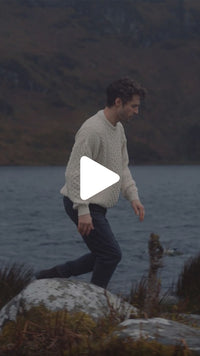 Person wearing a traditional Irish Aran crew neck sweater walking along a rocky lakeshore, with water and mountains in the background.