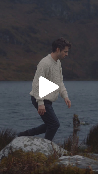 Person wearing a traditional Irish Aran crew neck sweater walking along a rocky lakeshore, with water and mountains in the background.