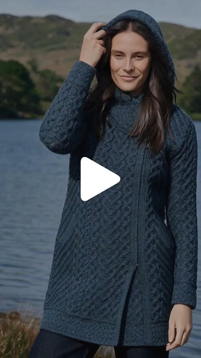 Video showcasing the Women's Heritage Aran Hooded Zipper Coat in blue, featuring traditional Irish cable knit patterns, a full-length zip closure, and a cozy hood, worn outdoors by a lakeside landscape.