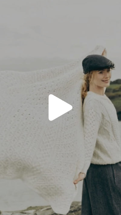 Person standing outdoors holding a large cream‑colored knitted wool throw draped over the shoulders, with a rocky coastal landscape in the background.
