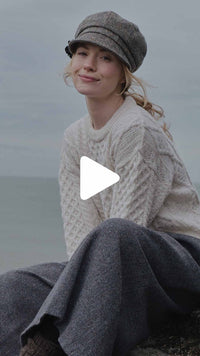Video showcasing a woman wearing an authentic white Aran cable knit sweater and a grey herringbone tweed newsboy cap in a coastal setting.