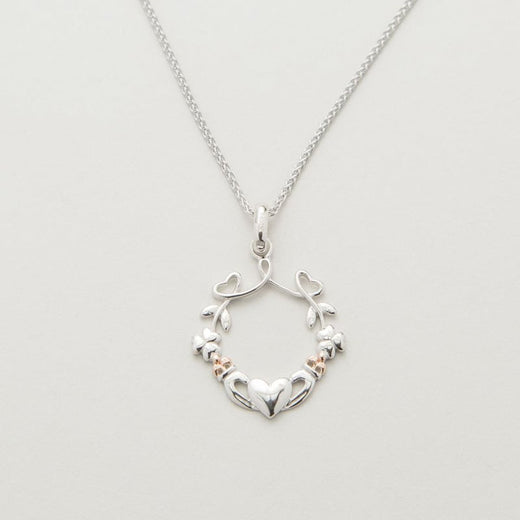 A silver pendant featuring a heart, shamrock, and claddagh design with rose gold accents on a chain.