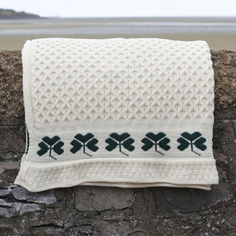 Cream-colored knit Aran blanket with textured diamond pattern and green shamrock border, draped over a stone wall by the beach—blending Irish-inspired textile design with coastal outdoor styling for artisan home décor and lifestyle photography.