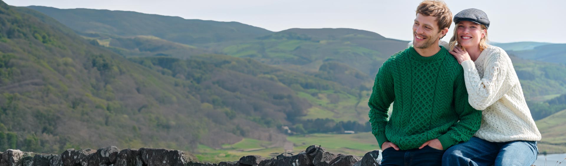 Two people wearing traditional aran sweater sitting on a stone wall with a scenic landscape in the background