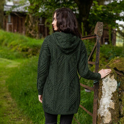 A woman with shoulder-length dark hair stands with her back to the camera, wearing a dark green Aran cardigan. She faces a grassy area with trees in the background and is holding onto a rustic gatepost. The setting appears serene and natural.