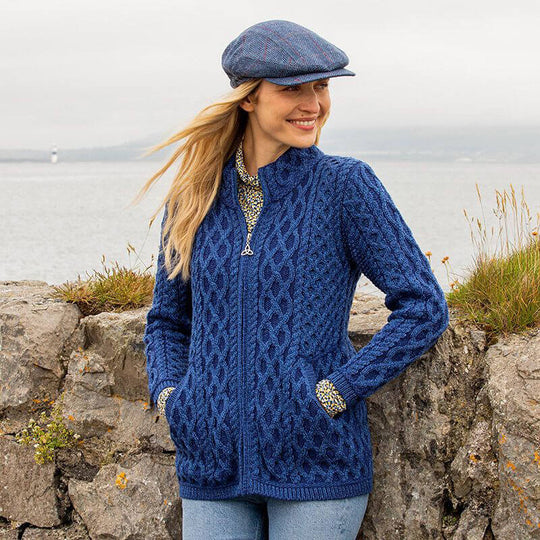 A woman stands next to a stone wall near the water, wearing The Women's Traditional Aran Zip-Front Cardigan in blue and a matching flat cap. Her long hair flows gently in the breeze as she smiles, with a scenic background of water and sky.