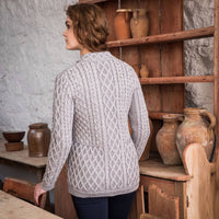 A woman with shoulder-length hair stands with her back to the camera, wearing a light gray, intricately knitted aran sweater. She is indoors, surrounded by rustic wooden furniture and clay pots on a shelf in the background. The setting has a cozy, homey feel.