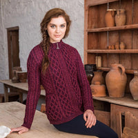 A young woman with long, wavy hair is wearing a dark burgundy cable-knit aran sweater. She sits at a wooden table in a rustic interior, surrounded by clay pots displayed on shelves. The setting features stone walls and wooden furniture, creating a cozy atmosphere.