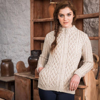 A woman stands in front of a rustic wooden shelf, wearing a cream-colored, cable-knit aran sweater with a high collar and pockets. Her hair is styled in a braid, and she poses confidently. The background features clay pots and wooden furniture, enhancing the cozy atmosphere.