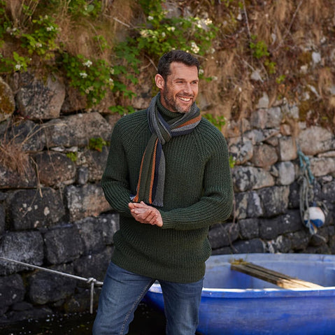 A man standing outdoors wearing a green knitted sweater and a striped scarf around his neck.