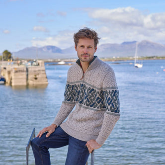 A man sitting on a rock wearing a green aran sweater with Celtic knot patterns, smiling at the camera with a body of water and landscape in the background.