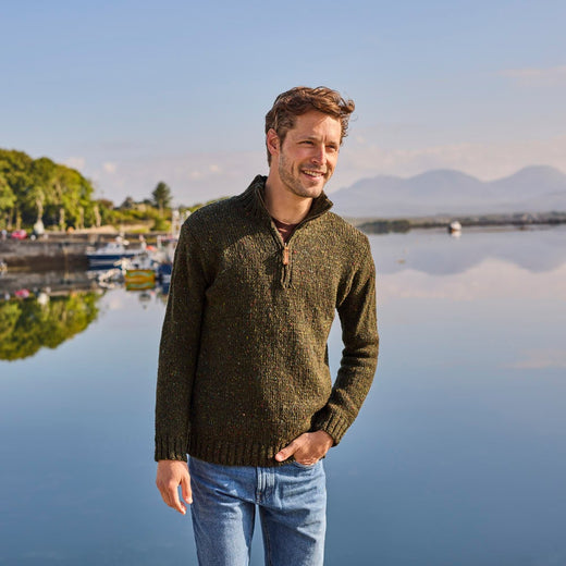 Man wearing a forest green men's half zip fleck sweater and blue jeans, standing near a calm waterfront with trees, boats, and distant mountains in the background