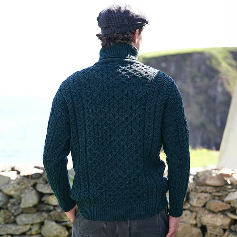 Back view of the Men's Signature Fisherman Turtleneck Wool Sweater in Blackwatch, showcasing intricate traditional Irish Aran stitching and a classic roll neck design, styled beside a traditional stone wall with dramatic Irish coastal cliffs in the background.
