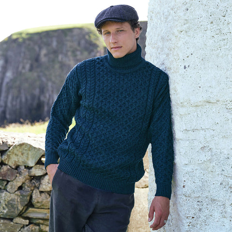 Men's Signature Fisherman Turtleneck Wool Sweater in Blackwatch, crafted from premium wool with traditional Irish Aran stitching and a classic roll neck design, styled beside a traditional stone cottage overlooking the rugged Irish coastline.