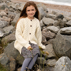 Girl sitting on a rocky beach waering a cream merino wool Kids  Aran cardigan with traditional cable knit patterns and brown buttons