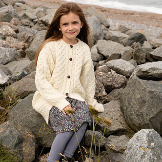 Girl sitting on a rocky beach waering a cream merino wool Kids  Aran cardigan with traditional cable knit patterns and brown buttons