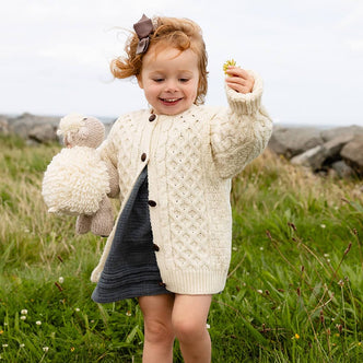 Child playing in a field wearing cream  kids Aran sweater with traditional Irish knitwear patterns. She holds a yellow flower and a teddy bear 