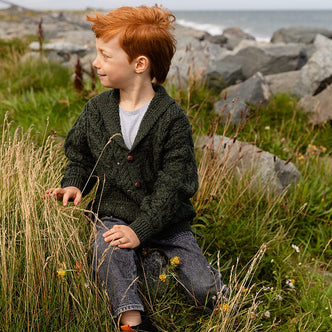 A young boy with red hair sits on a grassy hill, wearing a dark green kids aran cardigan and jeans. He looks thoughtfully to the side, with rocks and the ocean in the background. Wildflowers and tall grass surround him.