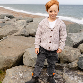 A young child stands on rocky terrain near the beach, smiling at the camera. The child has short red hair and is wearing a beige kid's aran cardigan over a white shirt, paired with dark jeans and black rubber boots. The ocean is visible in the background under a cloudy sky.