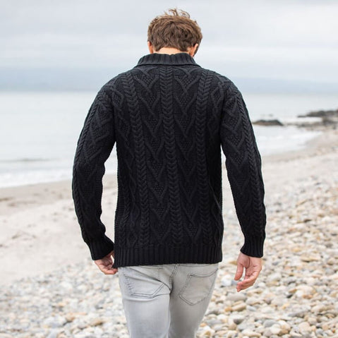 A man walking along a pebbled beach, viewed from behind. He is wearing a black, cable-knit aran sweater and light-colored jeans. The ocean is visible in the background, with calm waves and a cloudy sky.