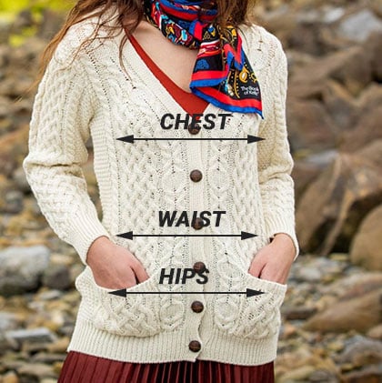 A woman stands outdoors, wearing a cream-colored aran cardigan over a red top. She is holding her hands in the pockets of the cardigan. Labels indicate measurement points on her body: 
