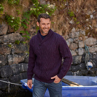 A man stands smiling near a stone wall and a small blue boat. He wears a dark purple, cable-knit aran sweater over blue jeans, with greenery partially visible in the background. The scene appears relaxed and outdoor-oriented.
