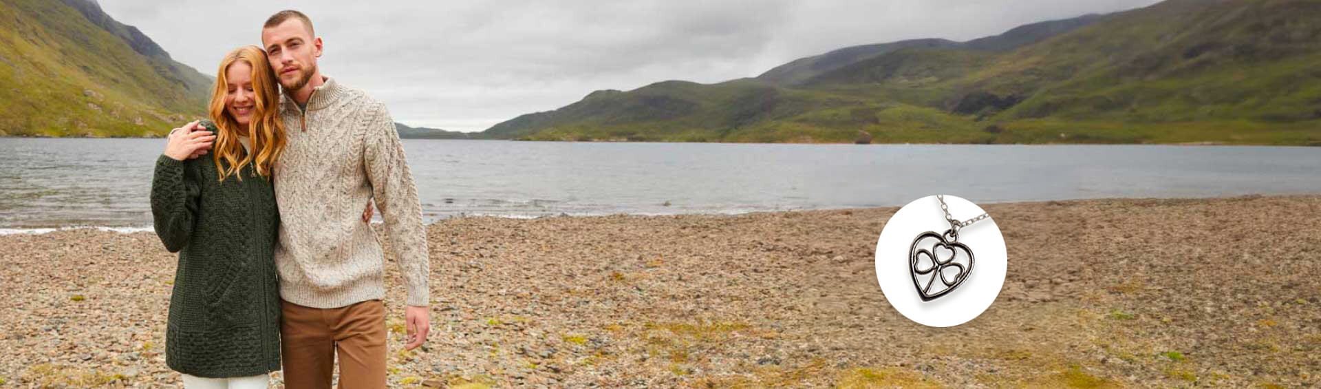Couple standing on a pebbly beach with mountains and water in the background, wearing matching knit sweaters.