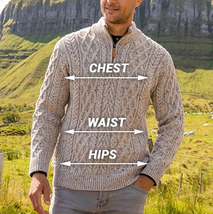 A man stands outdoors wearing a light-colored cable-knit aran sweater with a half-zip collar. The background features green hills and a rocky cliff. Labels indicate measurements for 