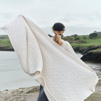 Traditional Aran honeycomb wool throw in natural cream with cable and diamond knit patterns, displayed outdoors near a scenic coastal landscape with green hills and water.