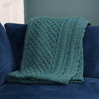 green cable-knit throw blanket with diamond lattice pattern draped over a navy blue couch, styled against a teal accent wall and white trim—perfect for cozy, modern living room décor and textile inspiration.