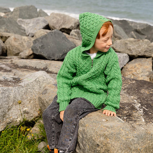 A child sits on a rock beside the water, wearing a bright green knitted  kids aran sweater and matching hood. The background features rocky terrain and a calm sea.