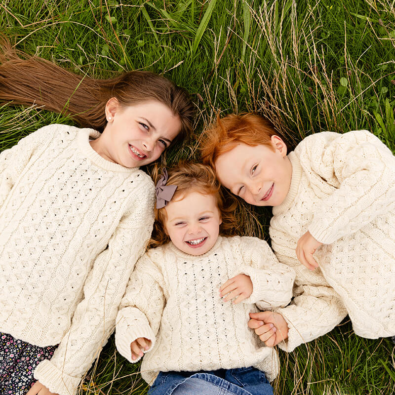 Three children lie on a grassy field, smiling at the camera. They wear cozy, matching cream-colored kids Aran sweaters. The middle child, a young girl with curly hair, grins widely while flanked by a boy with red hair and an older girl with dark hair.