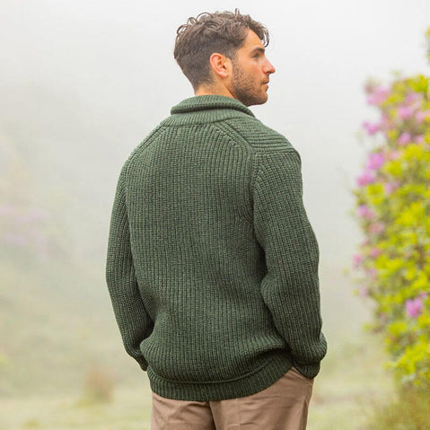 A man stands with his back to the camera, wearing a dark green knitted aran sweater. He has short, wavy hair and is looking into a misty landscape filled with soft colors and greenery. Pink flowers are visible in the background. The scene conveys a serene, natural atmosphere.