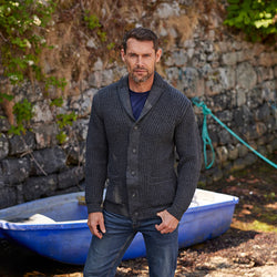 A man stands in front of a stone wall near a blue boat. He wears a dark gray, buttoned Aran cardigan over a navy shirt and has an attentive expression. The setting appears outdoors with some greenery in the background.