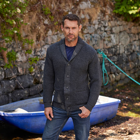 A man stands in front of a stone wall near a blue boat. He wears a dark gray, buttoned Aran cardigan over a navy shirt and has an attentive expression. The setting appears outdoors with some greenery in the background.