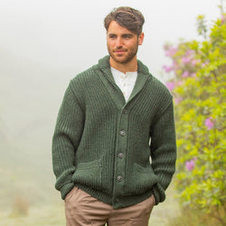 A man stands outdoors in a lush, misty environment, wearing a green knit cardigan over a white shirt. He has short brown hair and is looking slightly to the side, with purple flowers and greenery visible in the background.
